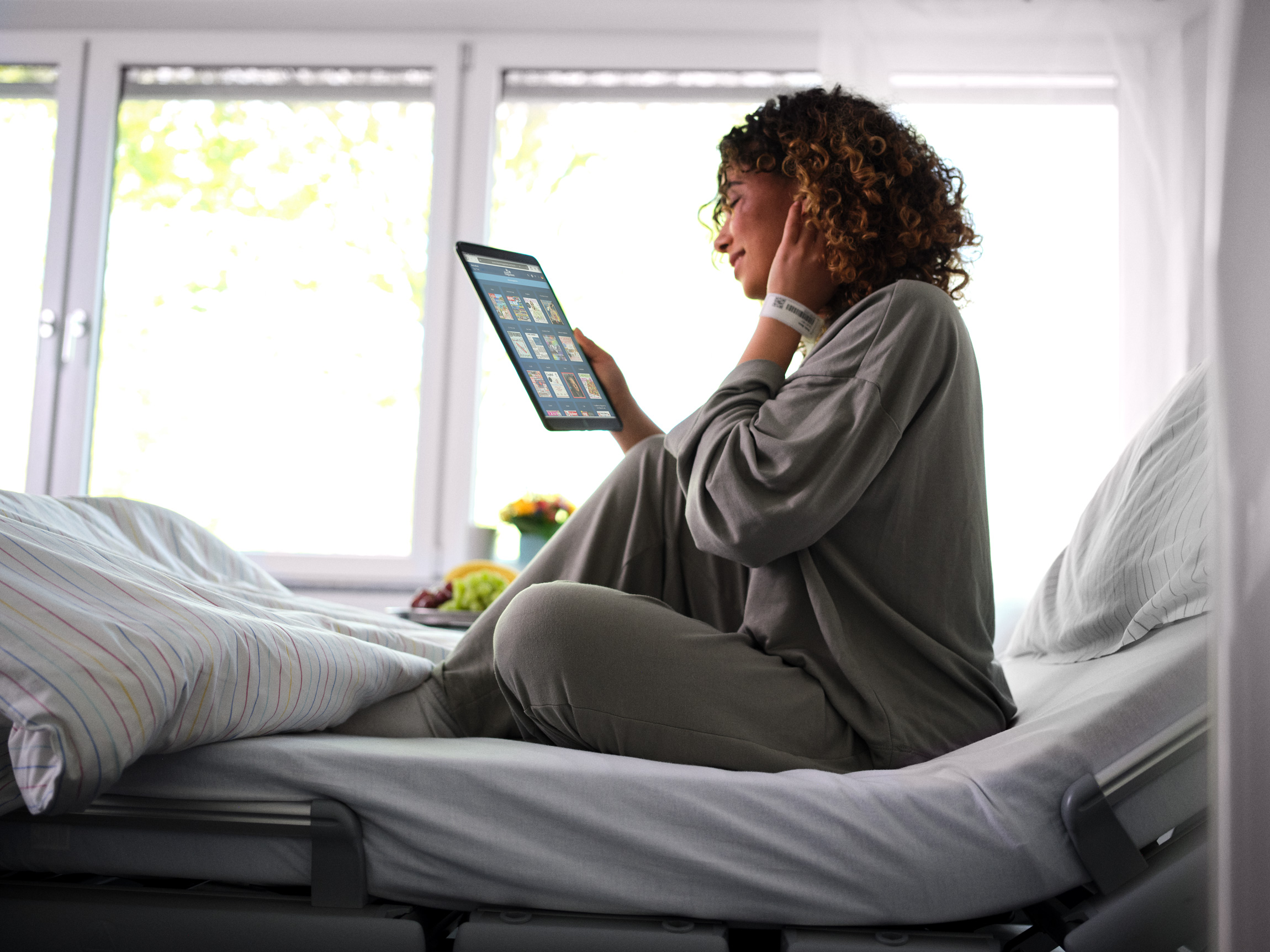 Patientenentertainment: Junge Patientin sitzt im Krankenhausbett und liest eMagazine von sharemagazines auf Tablet