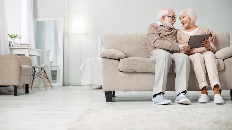 ConnectedCare Third Party Partner | GOLDENSUMMER.TV, Senioren-Ehepaar sitzt Arm in Arm auf Sofa im Wohnzimmer mit Tablet