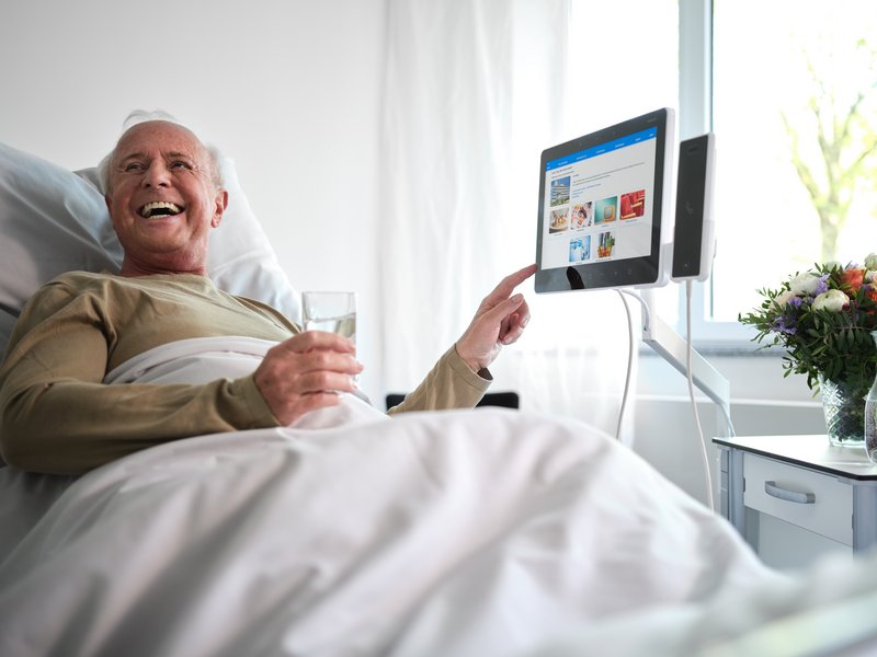 Älterer Patient im Krankenhausbett bedient lachend das ConnectedCare Tablet und hält ein Glas Wasser in der Hand