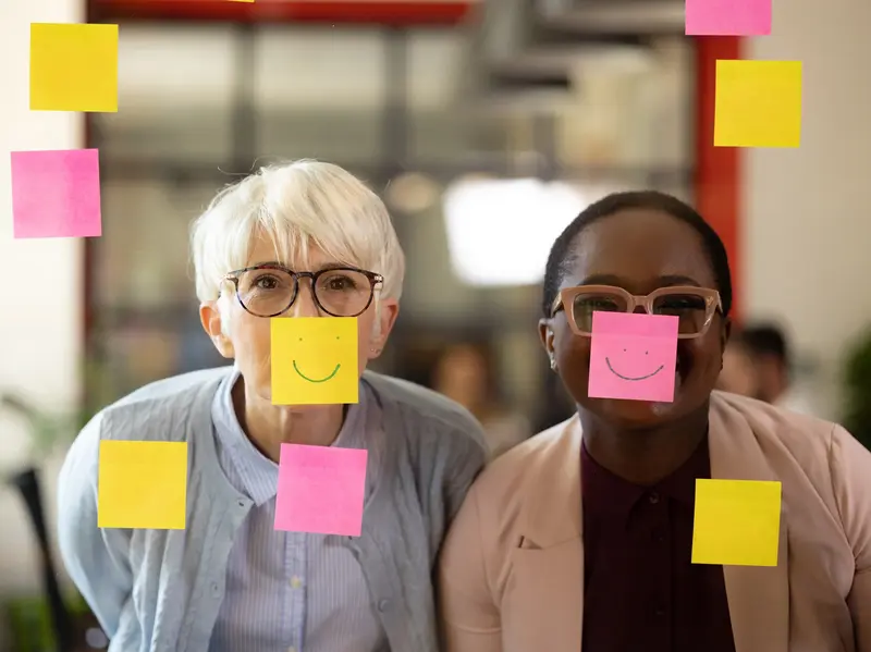 Ältere weiße Frau und junge schwarze Frau haben Spaß im Büro mit Post-Its auf Glasscheibe.