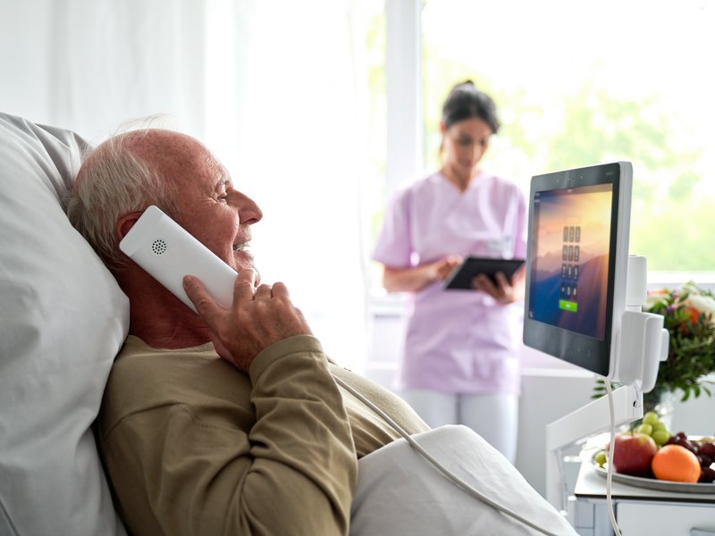 ConnectedCare Patientenkommunikation | Patient liegt im Bett und telefoniert mit ConnectedCare Phone, Pflegerin im Hintergrund