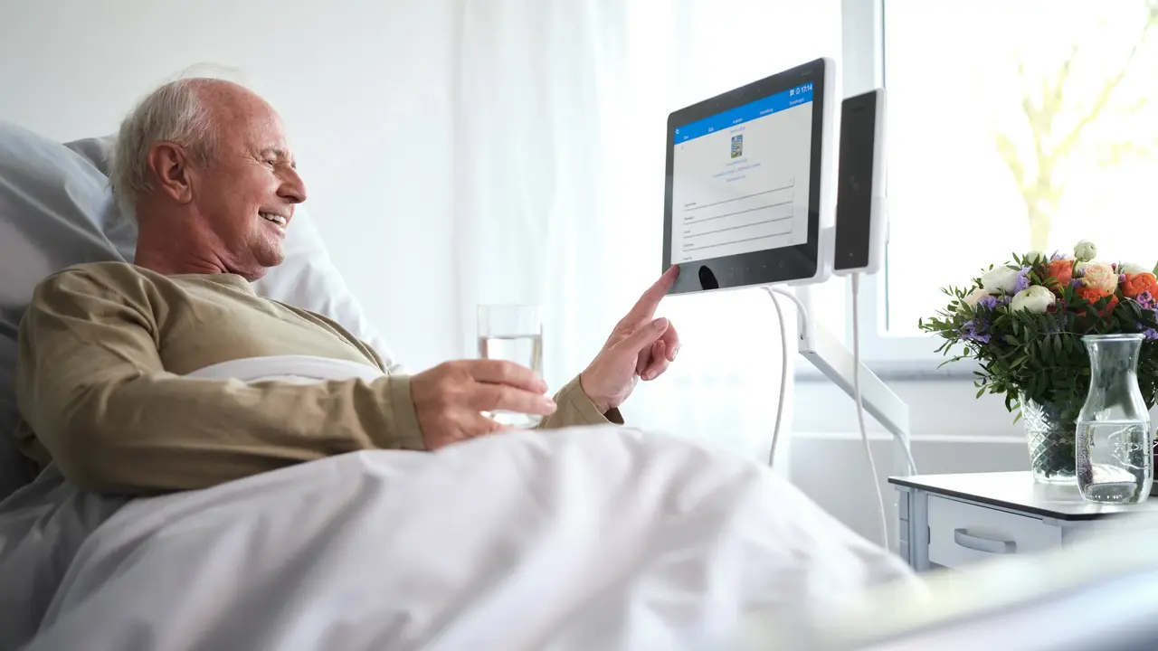 Patient Engagement | Älterer Patient liegt im Krankenhausbett und bedient Bedside Terminal