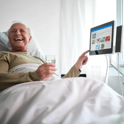 Älterer Patient im Krankenhausbett bedient lachend das ConnectedCare Tablet und hält ein Glas Wasser in der Hand