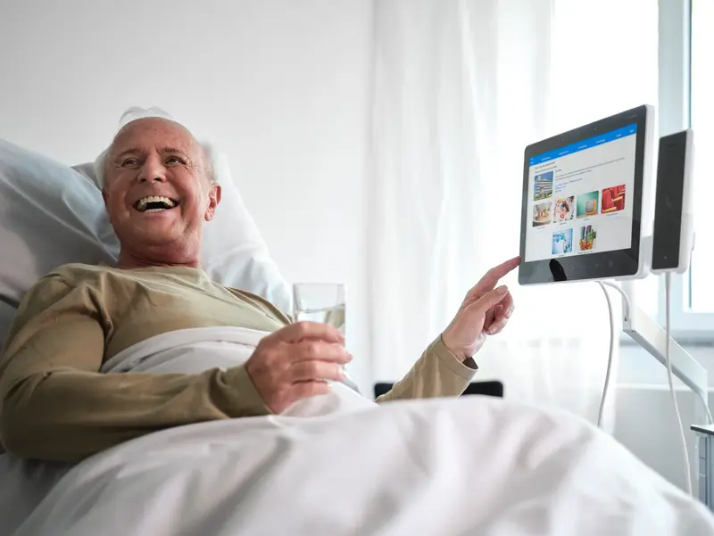 Älterer Patient im Krankenhausbett bedient lachend das ConnectedCare Tablet und hält ein Glas Wasser in der Hand