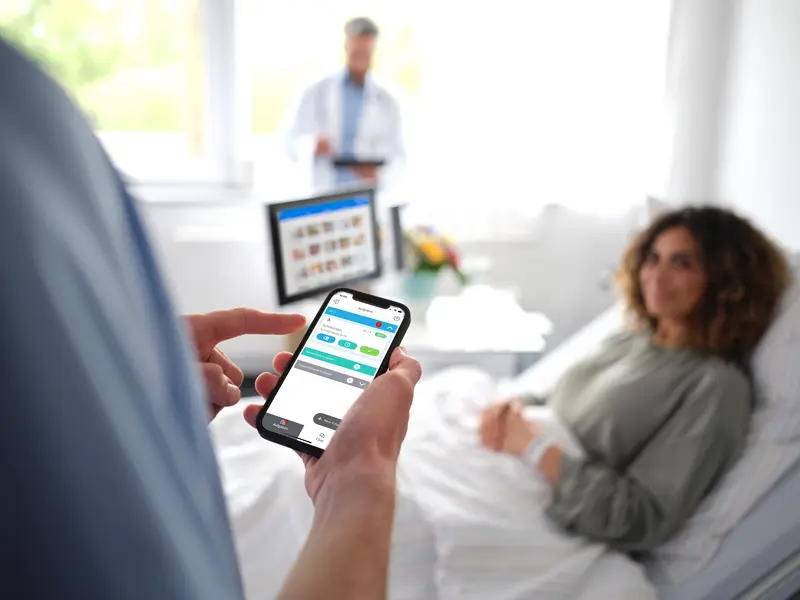 Digitale Patientenkommunikation: Pfleger steht am Patientenbett einer jungen Frau und bedient Smartphone mit dem Digitalen Serviceruf.