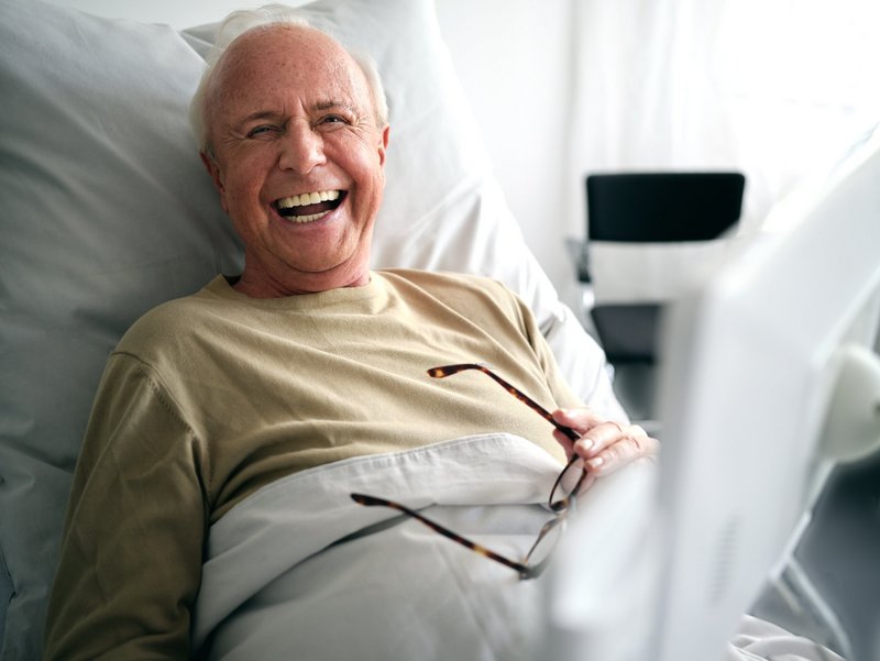 Älterer Patient im Krankenhausbett lacht herzhaft, ConnectedCare Tablet unscharf im Vordergrund