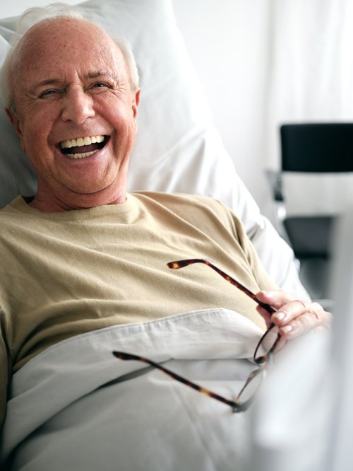 Älterer Patient im Krankenhausbett lacht herzhaft, ConnectedCare Tablet unscharf im Vordergrund