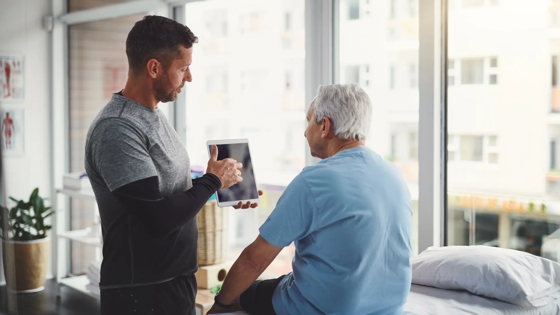 Physiotherapeut in Reha-Klinik zeigt älterem Reha-Patienten etwas auf einem Tablet.