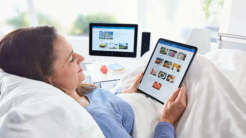 Patientin liegt im Krankenhausbett und schaut lächelnd auf Tablet mit ConnectedCare.